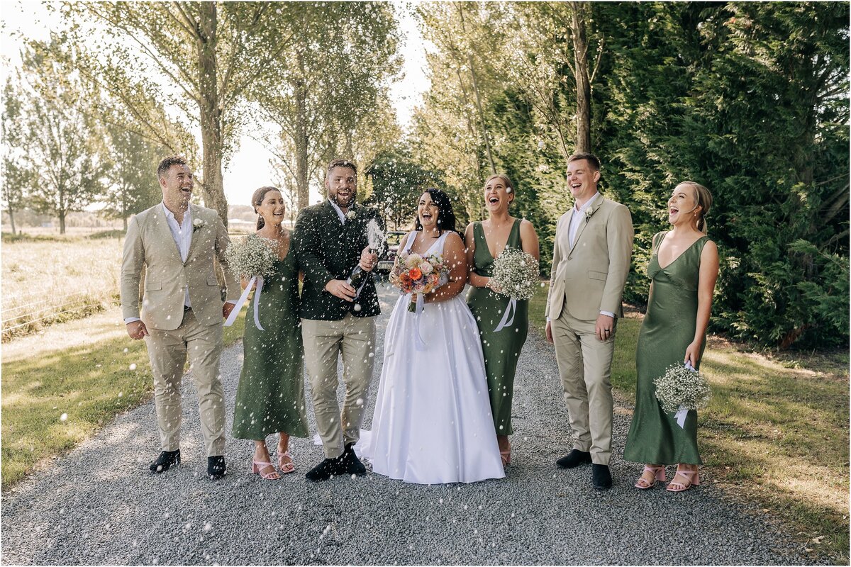 bridal party shaking champagne in driveway christchurch ohoka green trees loving ellies belly wedding green dresses laughing