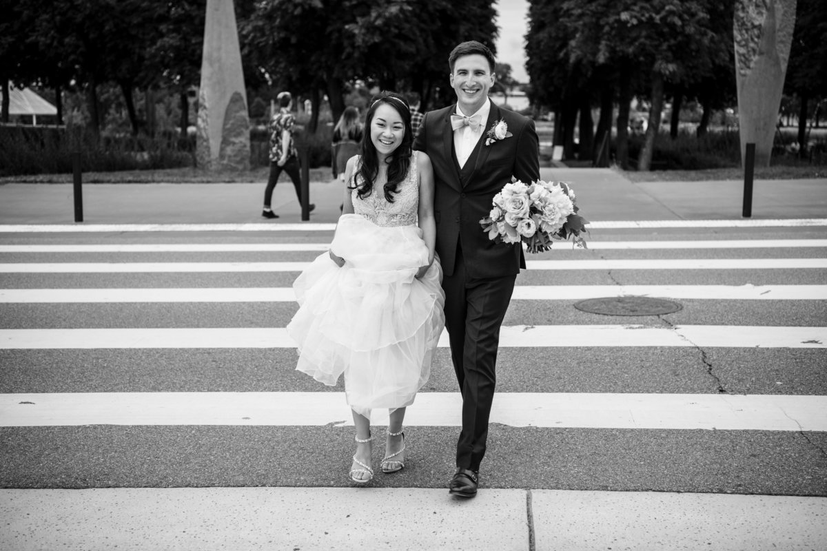 Asian bride and groom smile while walking across the street.