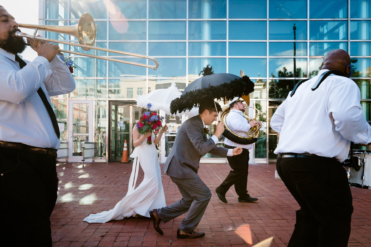 A bride and groom walking along a brick plaza with umbrellas as a band plays around them.