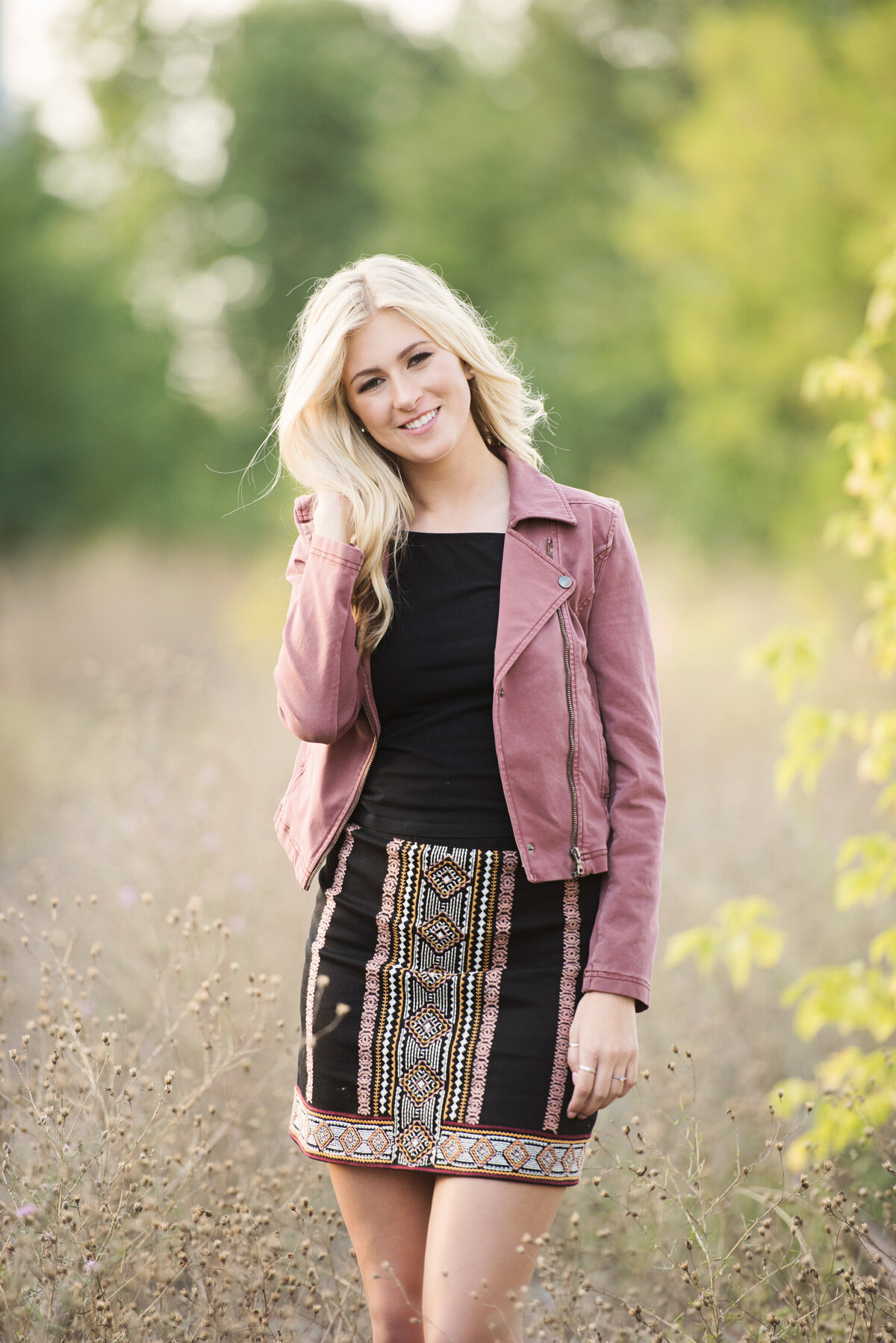 high school senior girl wearing pink jacket and patterned skirt in long grasses