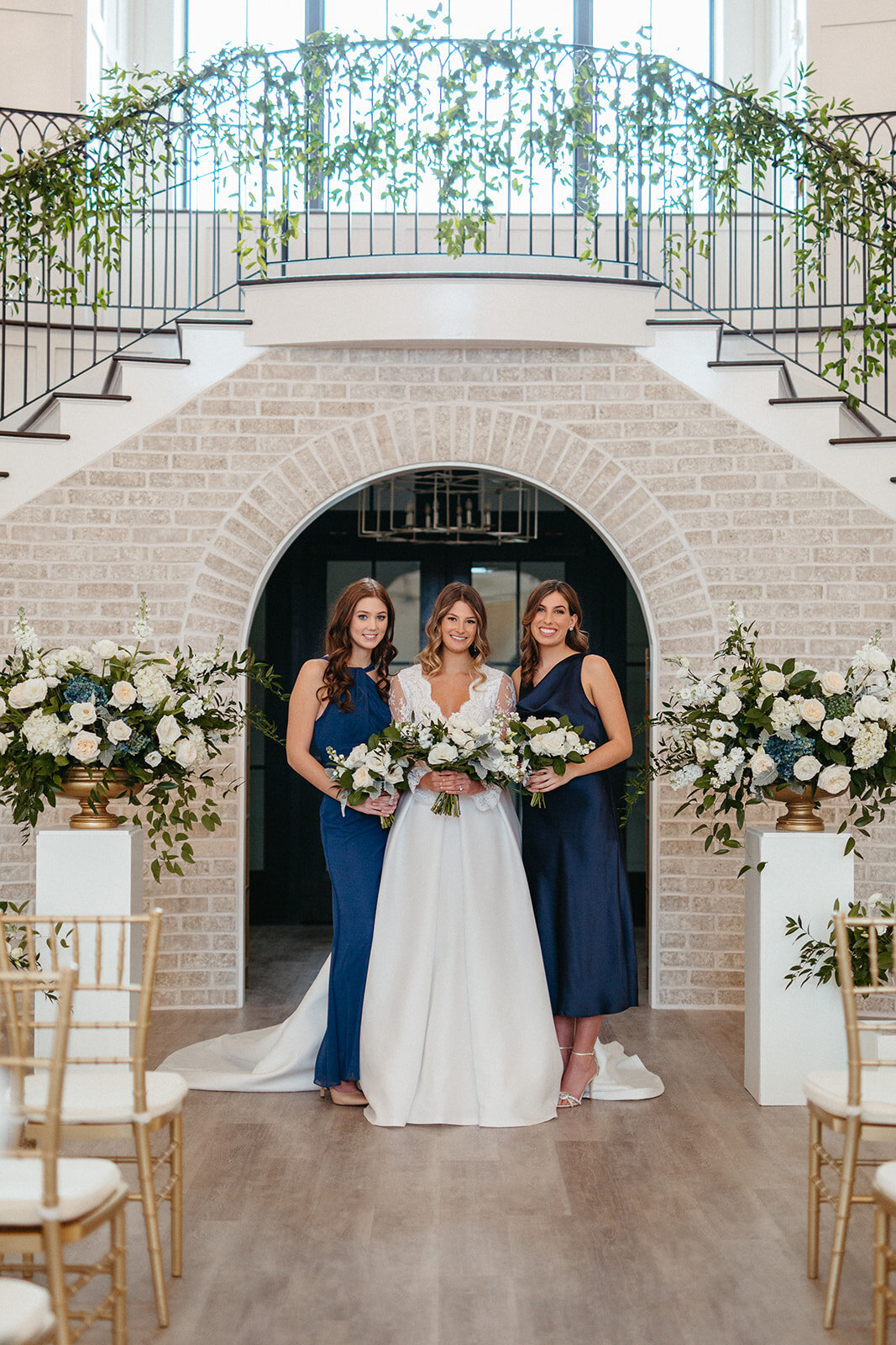 A bride in a white wedding gown holds a bouquet with two bridesmaids in blue satin gowns in front of a brick arched wall.