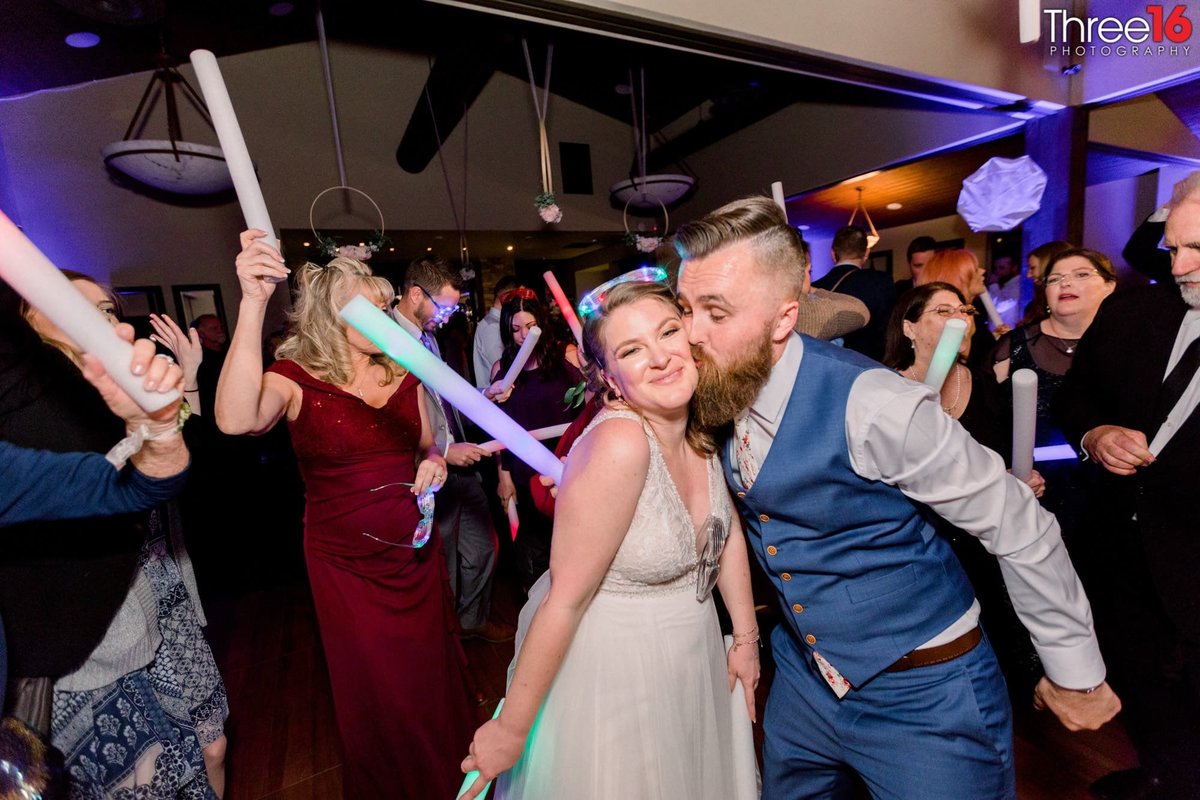 Groom kisses his Bride on the cheek while dancing with their wedding guests