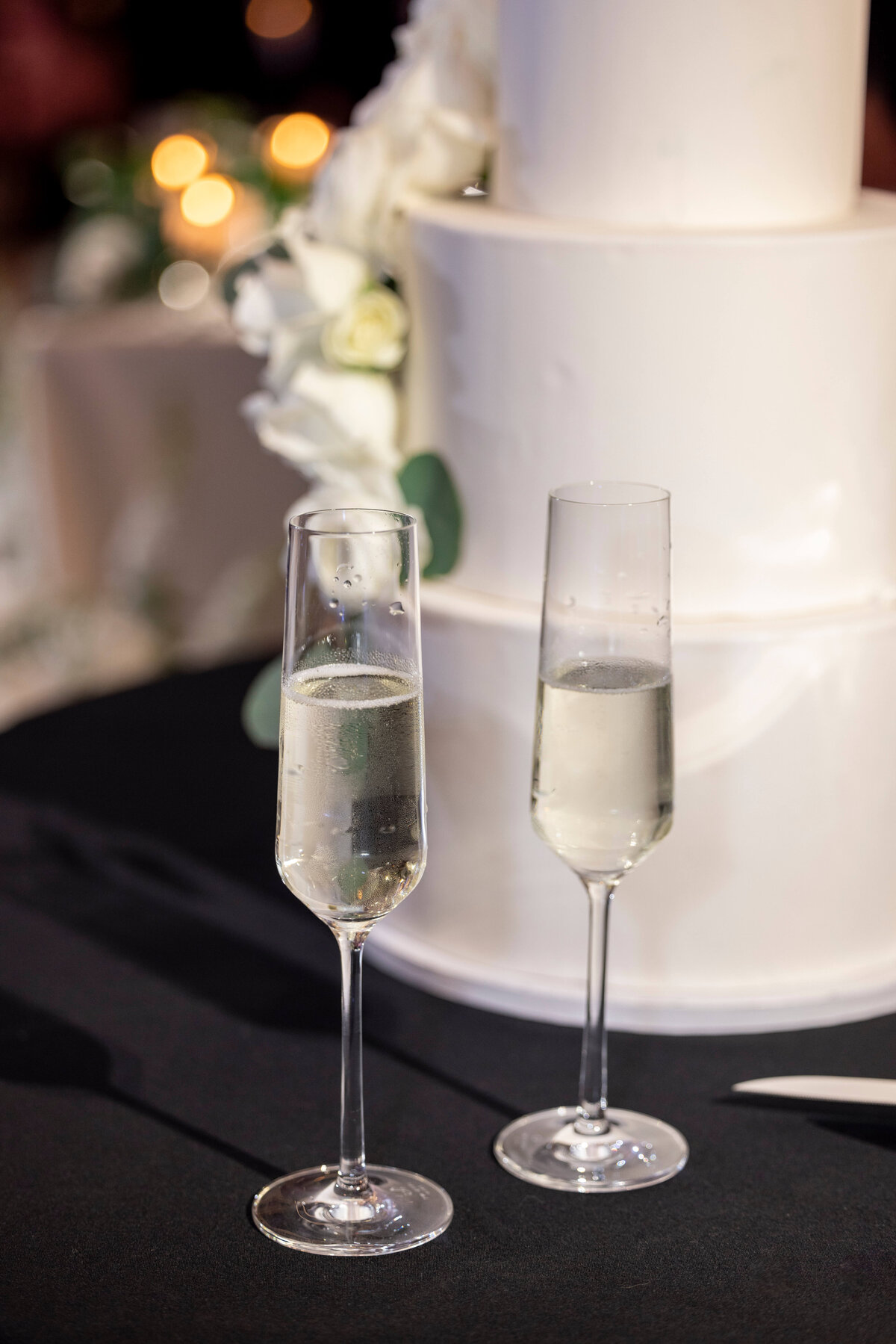 Two champagne flutes filled with bubbly liquid are placed on a table in front of a white, tiered wedding cake adorned with white flowers. The background has soft, out-of-focus lights.