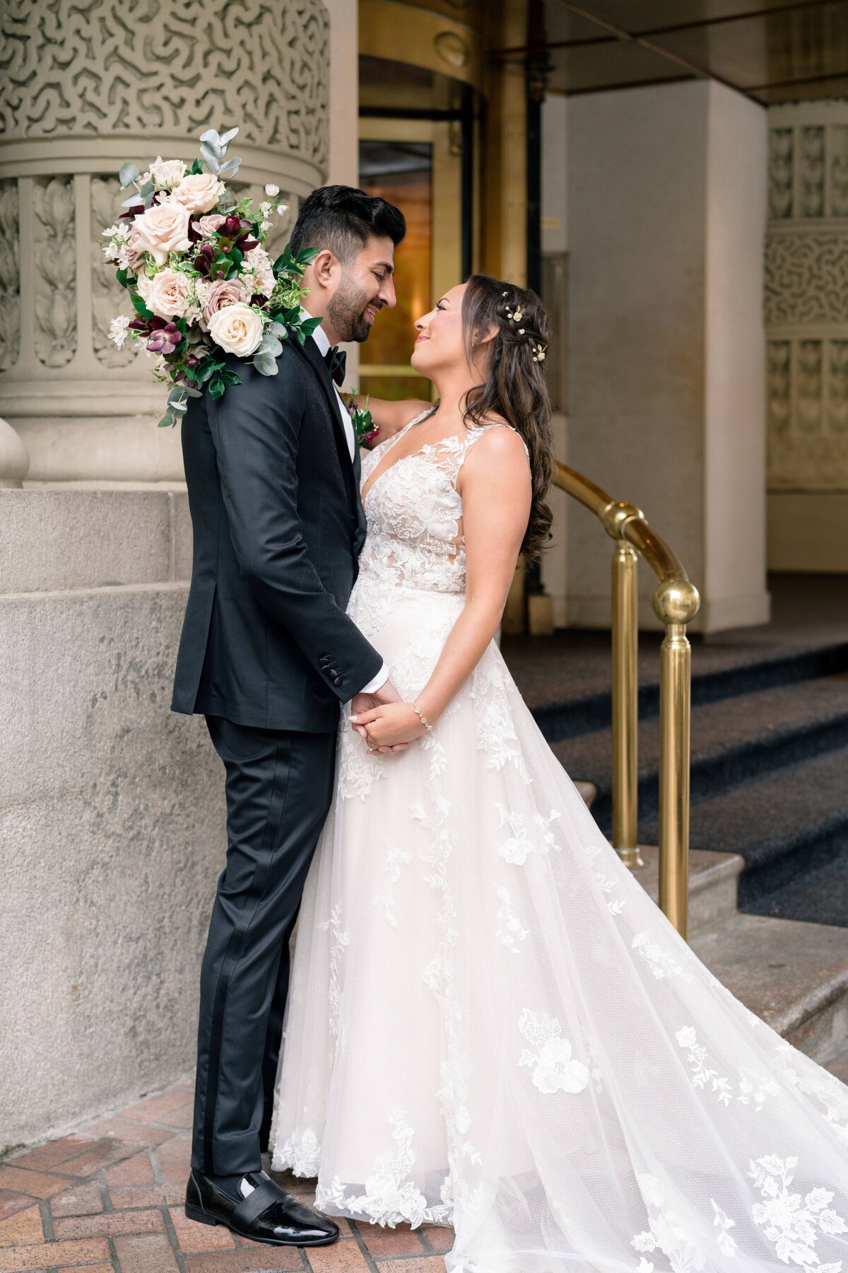 A bride and groom stand facing each other, holding hands and smiling. The bride wears a white lace gown and the groom is in a black suit. They stand outside a building with ornate details and a bouquet of flowers over the grooms shoulder.