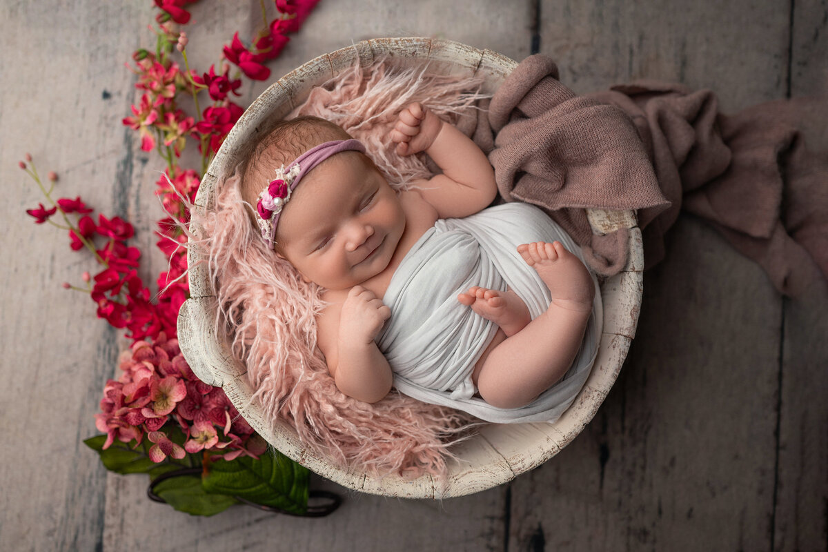 Portrait of sleeping infant in a wooden bucket swaddled in white cotton with arms and legs out. Baby girl is wearing   a floral headband and is surrounded by pink and red florals.