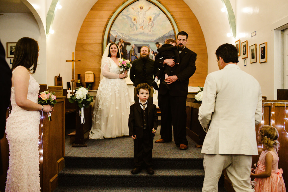 ring bearer stands in the middle of chapel in seward with chaos around