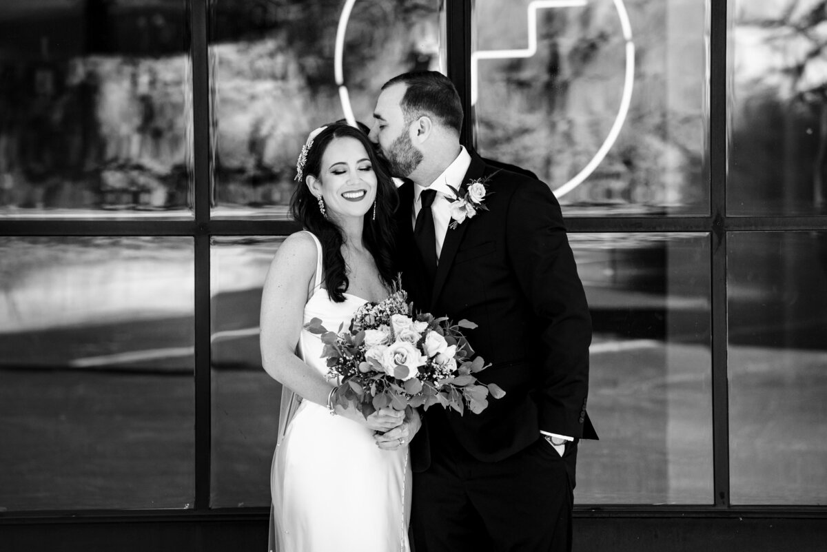 Black-and-white-image-of-groom-kissing-his-bride's-temple-as-she-smiles-and-closes-her-eyes-outside-the-doors-of-Upstairs-Atlanta