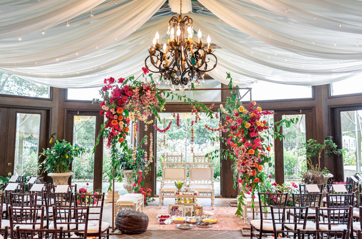 A beautifully decorated wedding ceremony setup with a floral arch, chairs arranged in rows, and a chandelier hanging from a draped ceiling. The large windows offer a view of the garden outside, enhancing the elegant ambiance.