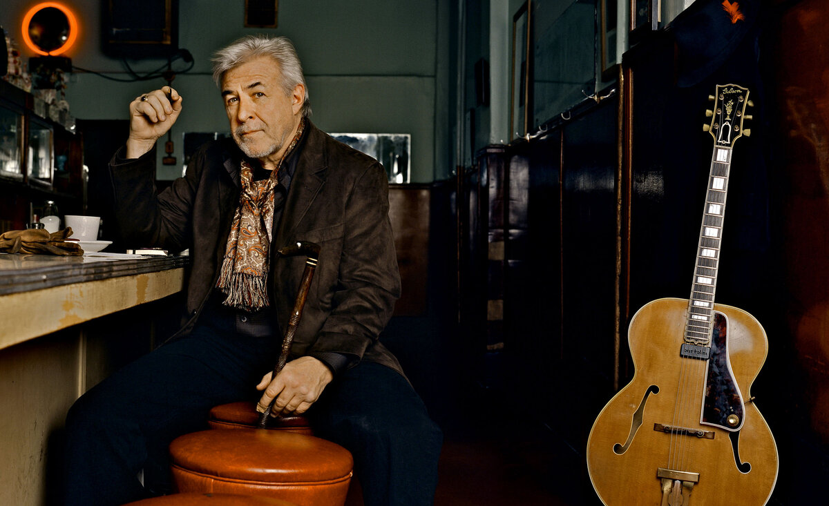 Male musician portrait Jim Byrnes wearing brown coat holding cain sitting against diner counter pale wood electric guitar beside