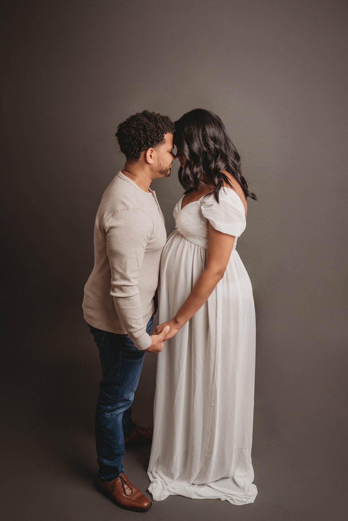 Man and woman 36 weeks pregnant facing each other, foreheads touching and holding hands. Woman is wearing white sheer maternity dress and man is wearing a cream long sleeve shirt with jeans and brown shoes.