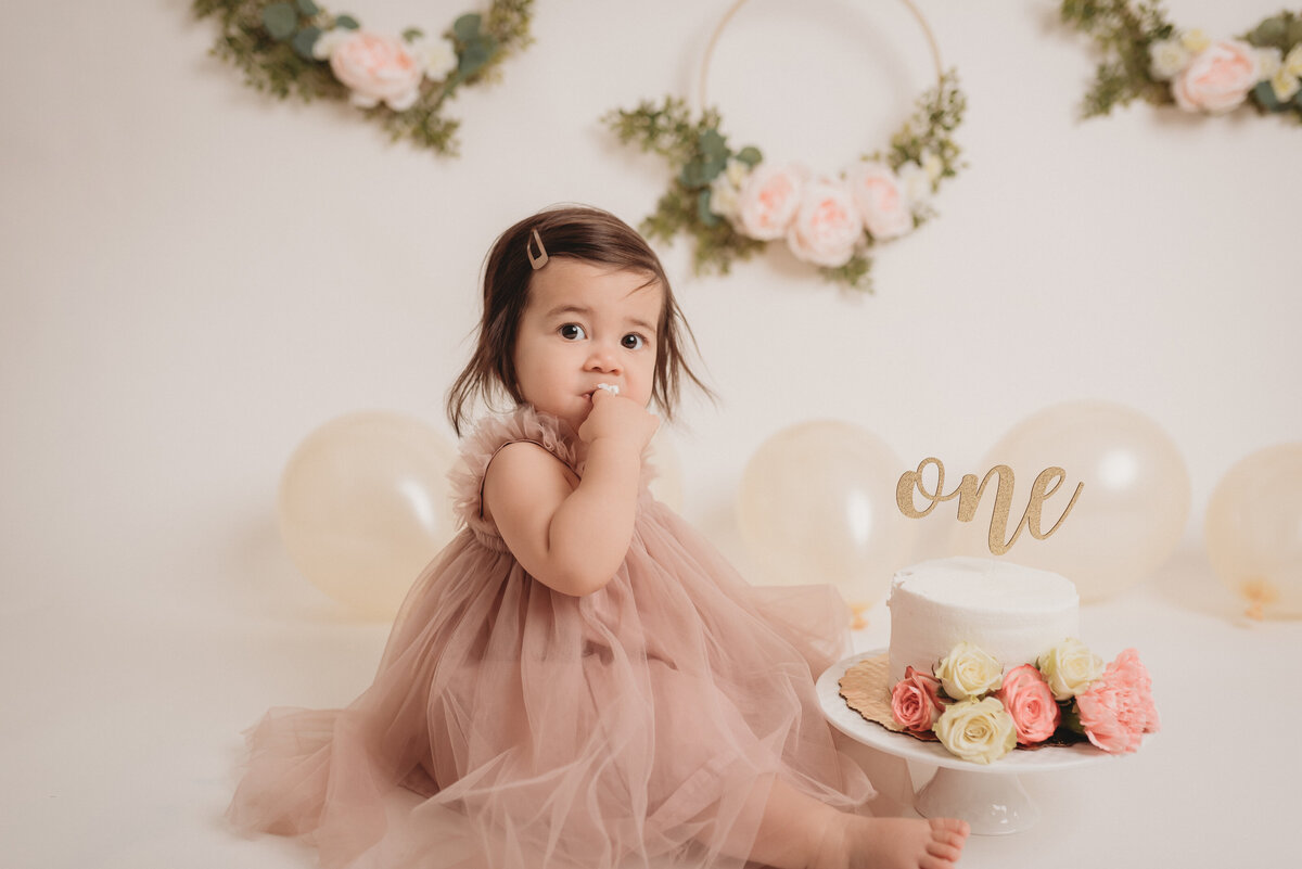 Cake smash portrait session at Atlanta photography studio with one year old girl in pink tulle dress eating cake with flowers on it and balloons and flowers in background