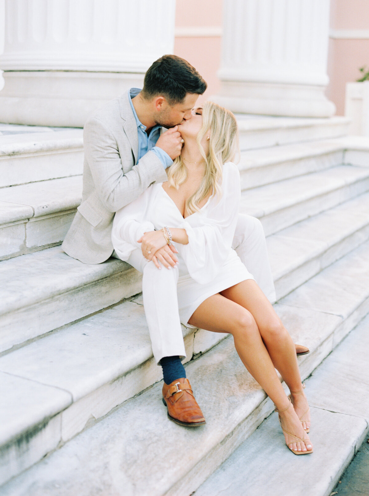 Downtown Charleston engagement session. Girl sitting in between guys legs on marble steps and kissing.