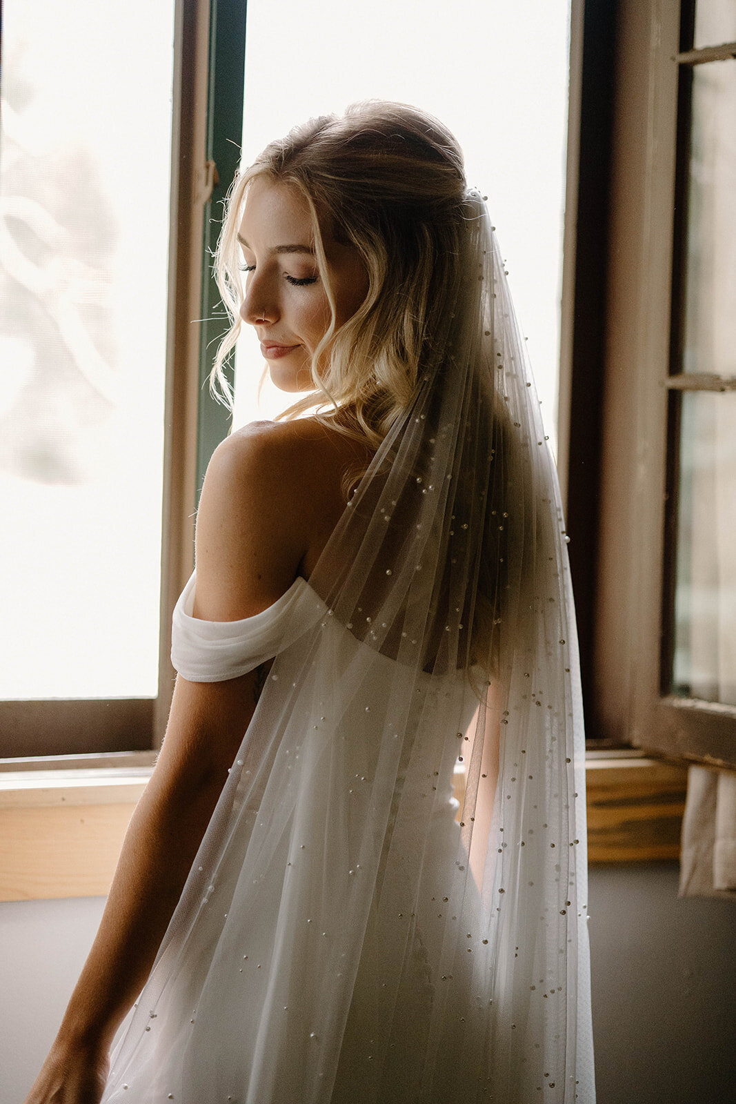 Stunning bride looks over her shoulder in bridal portrait framed in front of open window.