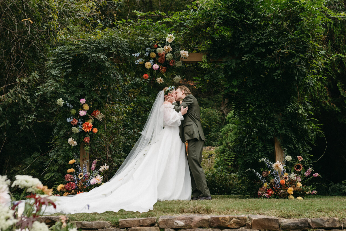 Bride and groom sharing their first kiss beneath a lush greenery arch accented with dahlias, roses, and wildflower-inspired floral clusters, creating a romantic garden ceremony scene at a Northwest Arkansas outdoor wedding.