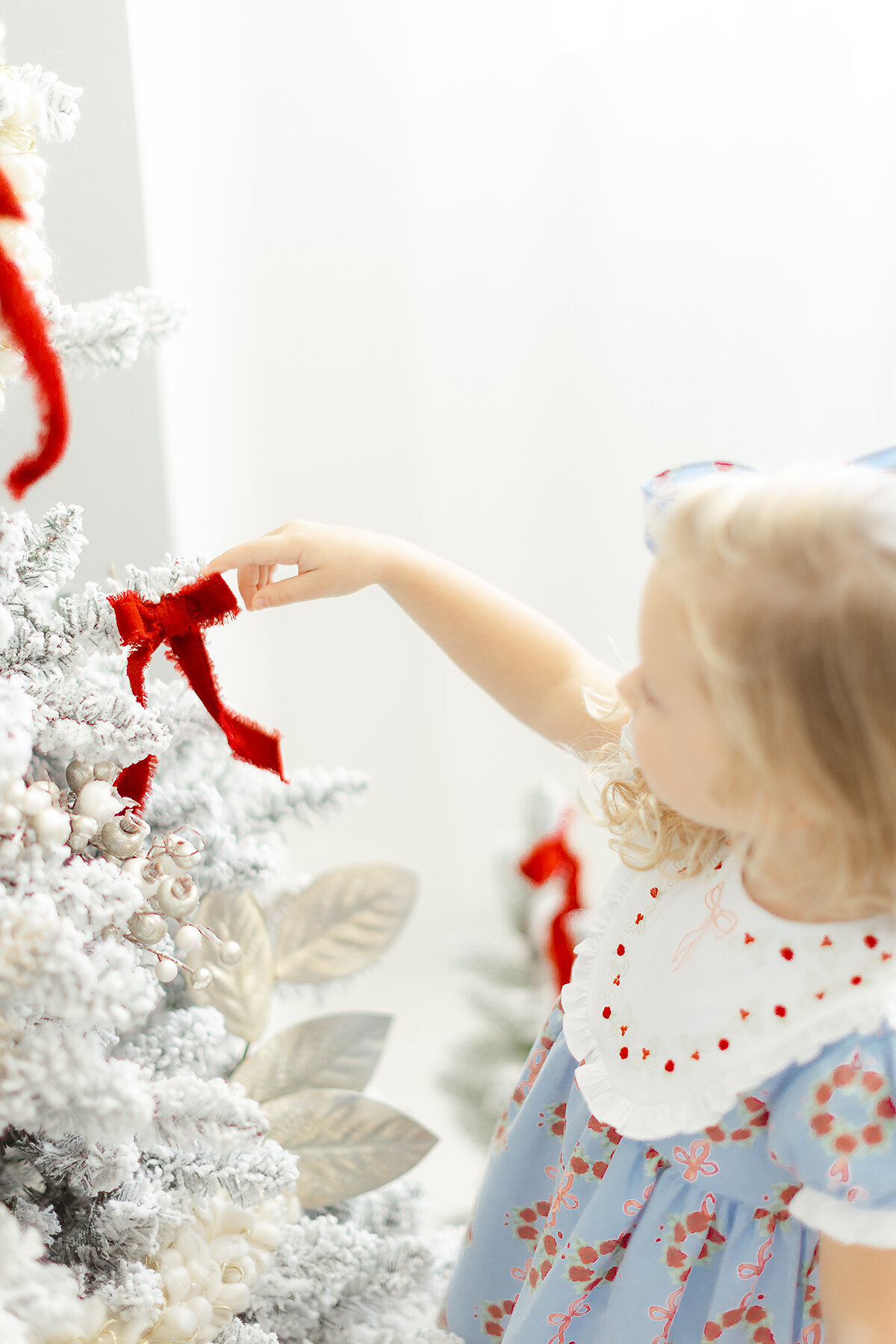 A little girl modeling a Dondolo christmas dress as she is touching a red velvet ribbon on a christmas tree.