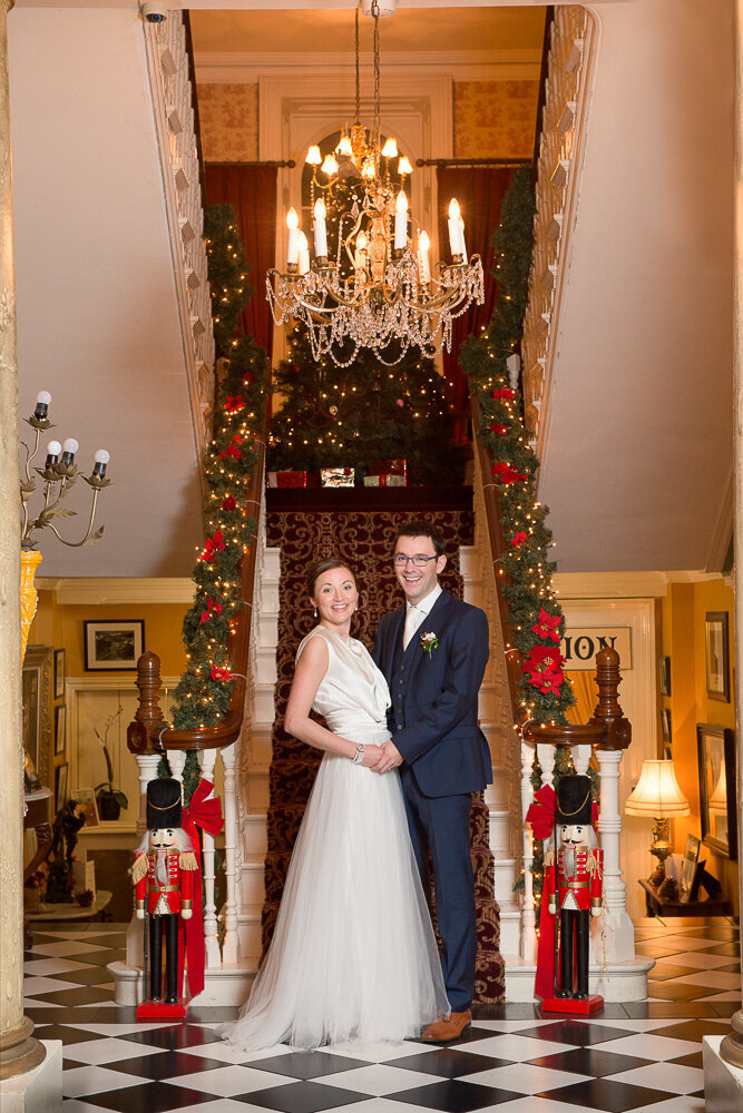 brunette bride wearing an a-line, tulle wedding dress with groom wearing a navy suit standing at the bottom of the stairs of Ballyseede Castle decorated for Christmas