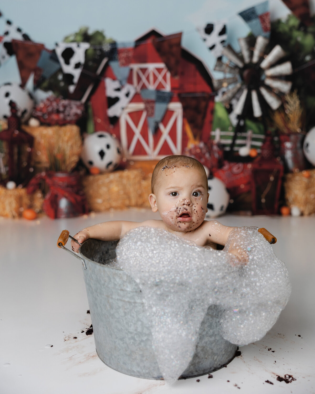 Baby boy cleaning up in a soapy tin tub with chocolate cake on his face, in front of a farm-themed first birthday setup. Photograph by Yucaipa cake smash photographer Kaitlyn Dawn Photography.