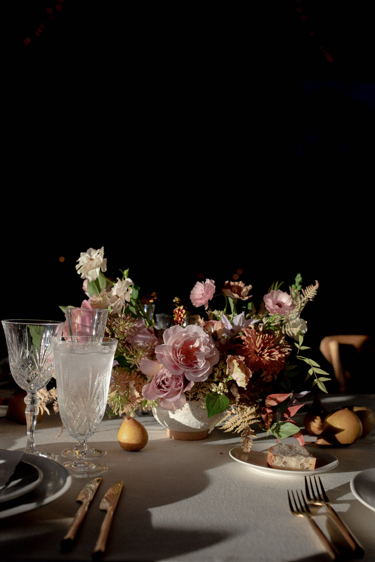 Dutch-inspired wedding centerpiece with gold flatware, crystal glasses, pears, and a bold bouquet with dramatic lighting.