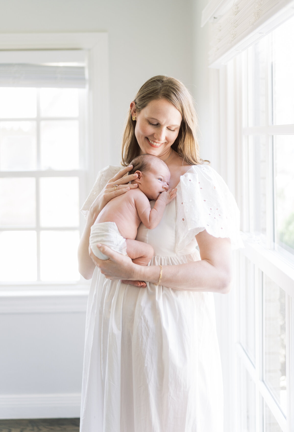 Portrait of a smiling woman in a white dress holding a newborn baby in her arms in front of a set of windows.
