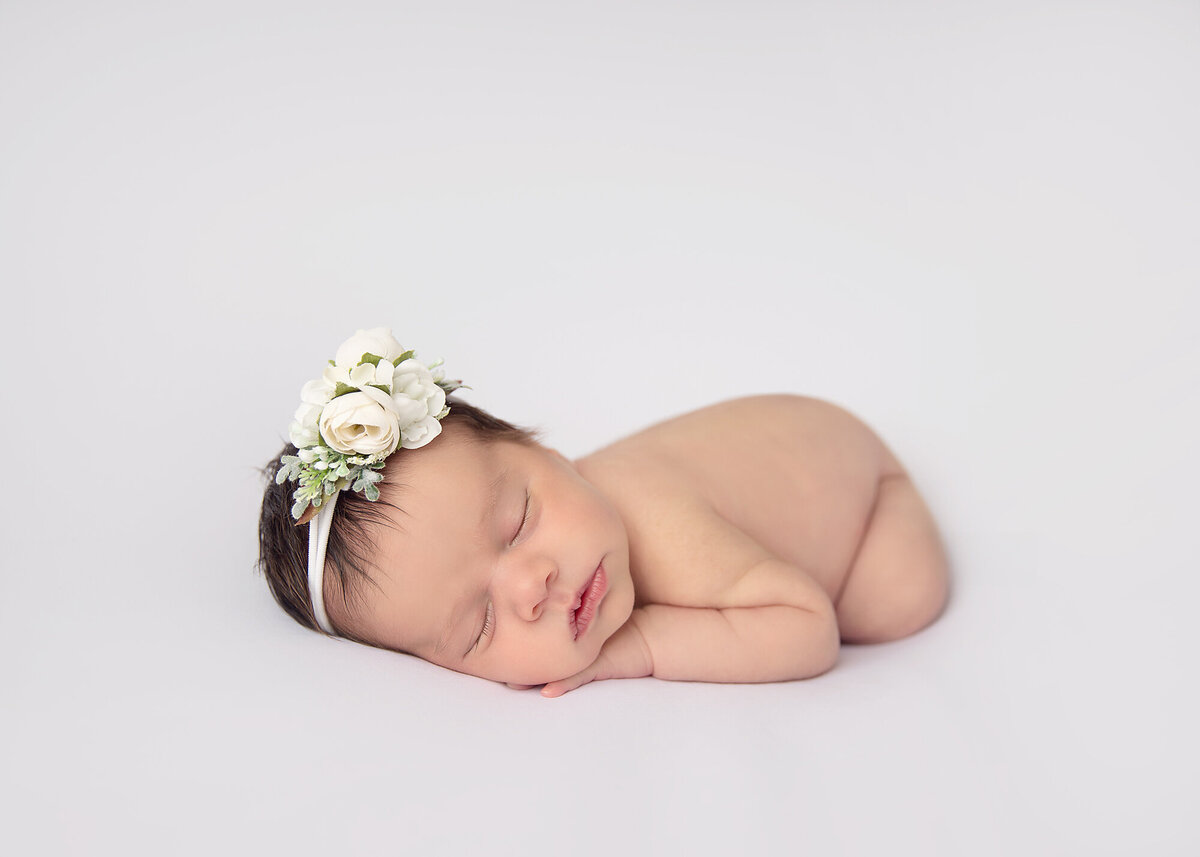 A newborn baby girl sleeps on her stomach in a studio with a white floral headband