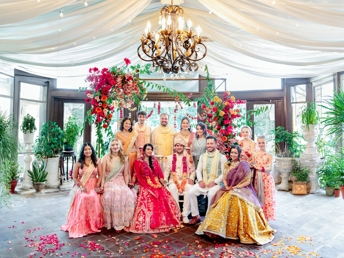 A vibrant cultural wedding scene with a couple seated in traditional attire, surrounded by a joyful wedding party. The setting features colorful outfits, floral arrangements, greenery, and a chandelier under a draped ceiling.