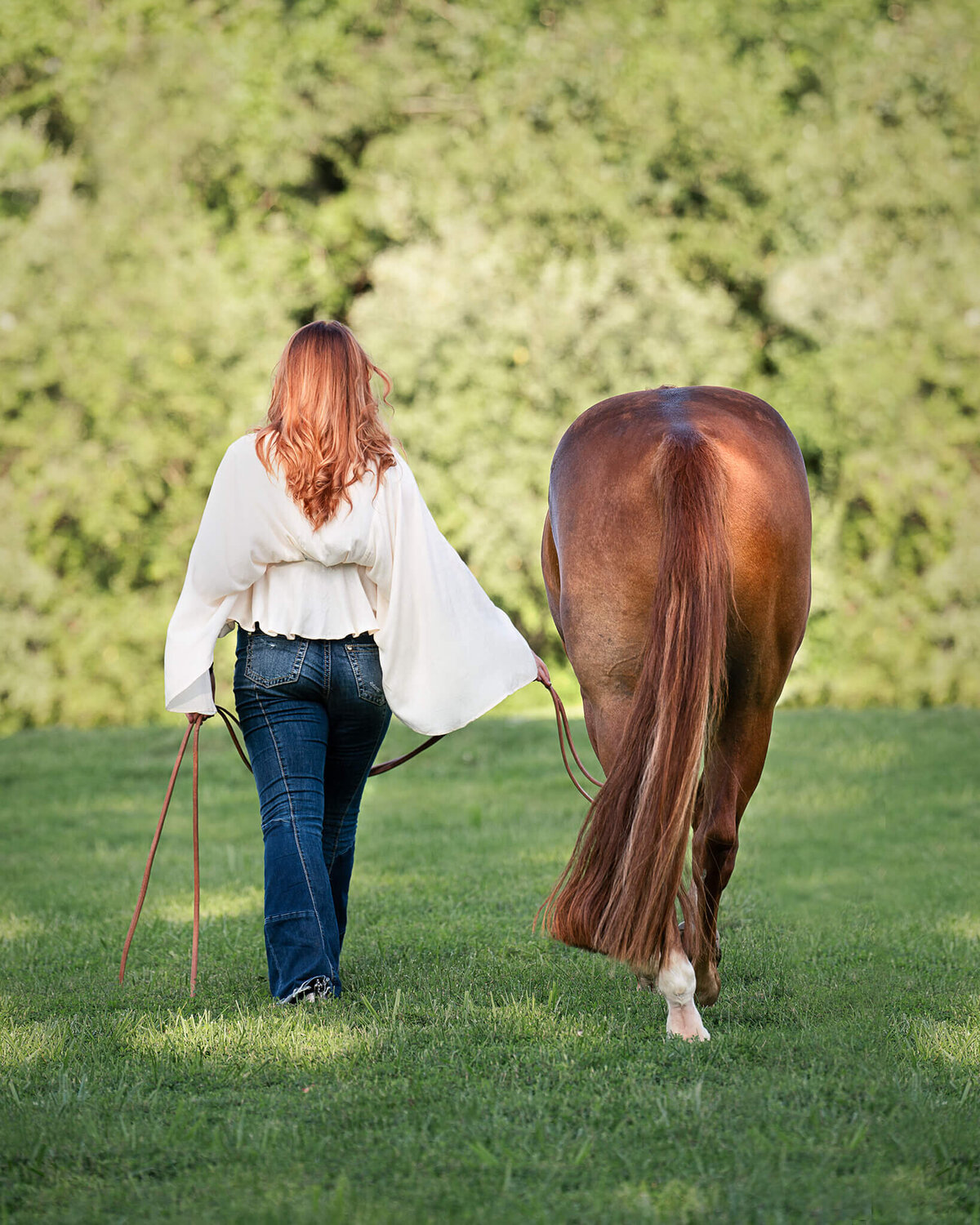 redhead equestrian female wearing a cream flowing shirt and jeans walking away from the camera with her quarter horse gelding by her side. They're walking away in a field with green grass surrounded by trees.