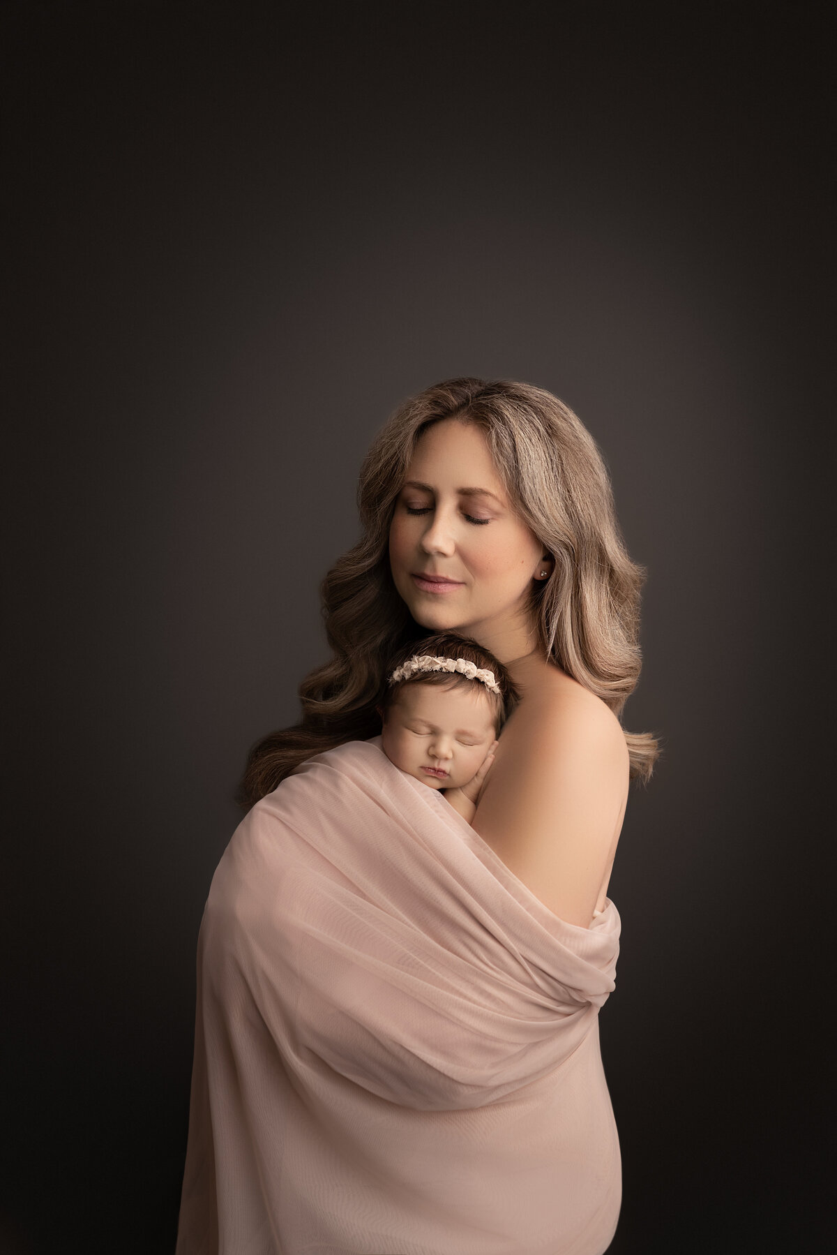 Fine art newborn photos captured by best Main Line newborn photographer Katie Marshall. Mom is holding her newborn baby girl, draped in a blush organza fabric. Baby girl is sleeping on mom's chest with one hand under her head. Mom's eyes are gently closed.