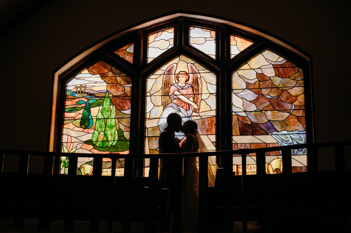 Photo of a bride and grooms silhouette in front of a stained glass window with an angel