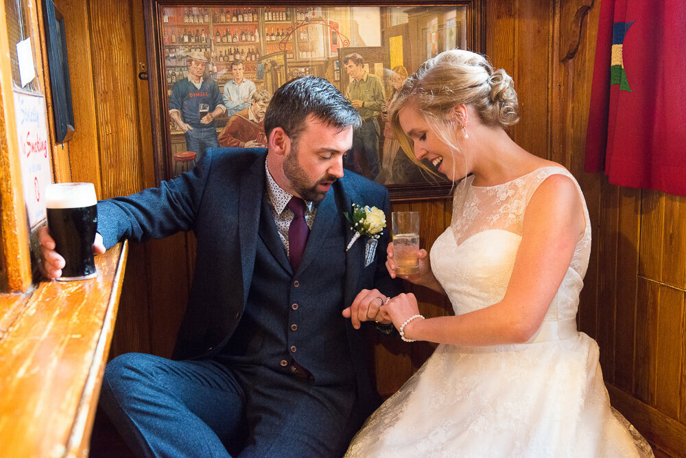 Bride with blonde hair, wearing a princess style wedding dress sitting in the snug in Dick Mack's pub with her groom, wearing a navy suit and waistcoat with wine tie, holding a Guinness and looking at the wedding rings