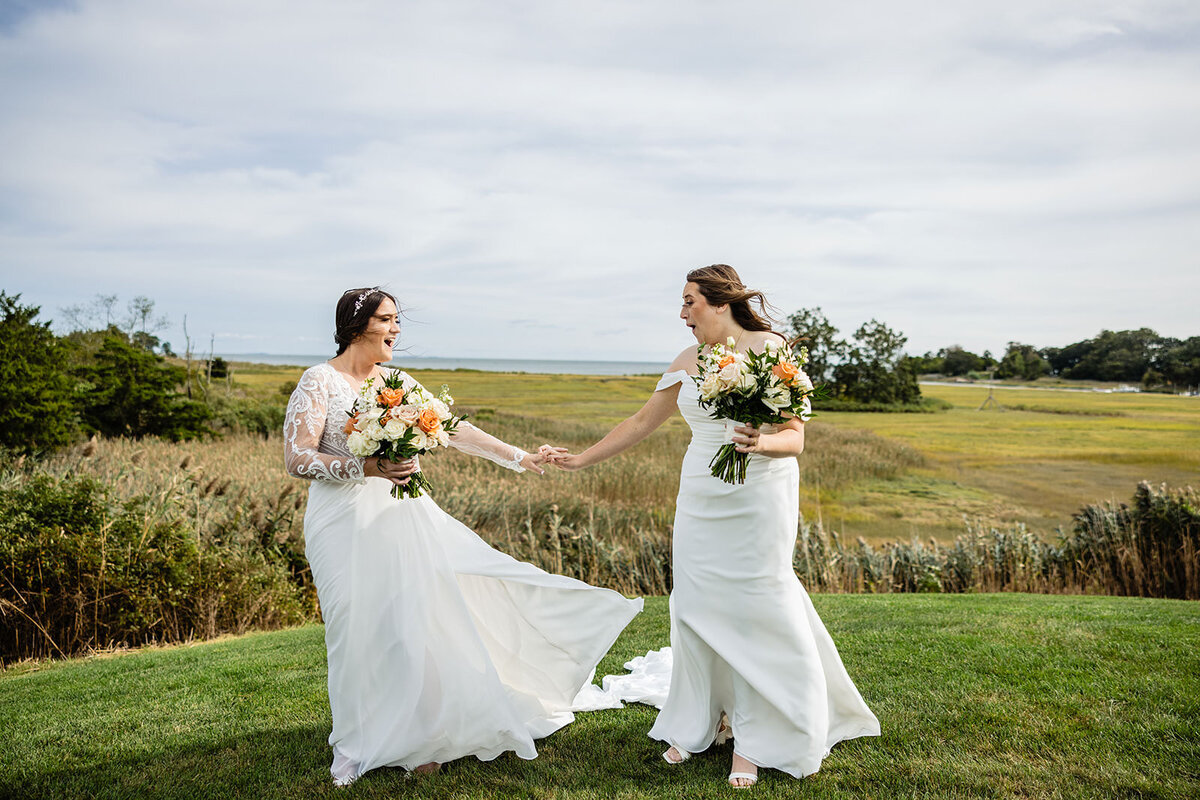 Two brides holding hands and looking at each other, standing in a field with a scenic view of a marshland behind them, both dressed in elegant wedding gowns and holding bouquets with orange flowers.