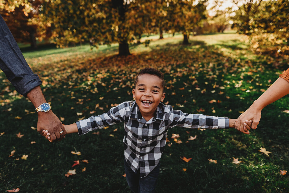 fall family photos with little boy holding hands with his parents as they play together in a park with fallen leaves on the green grass captured by Family photographers Maryland