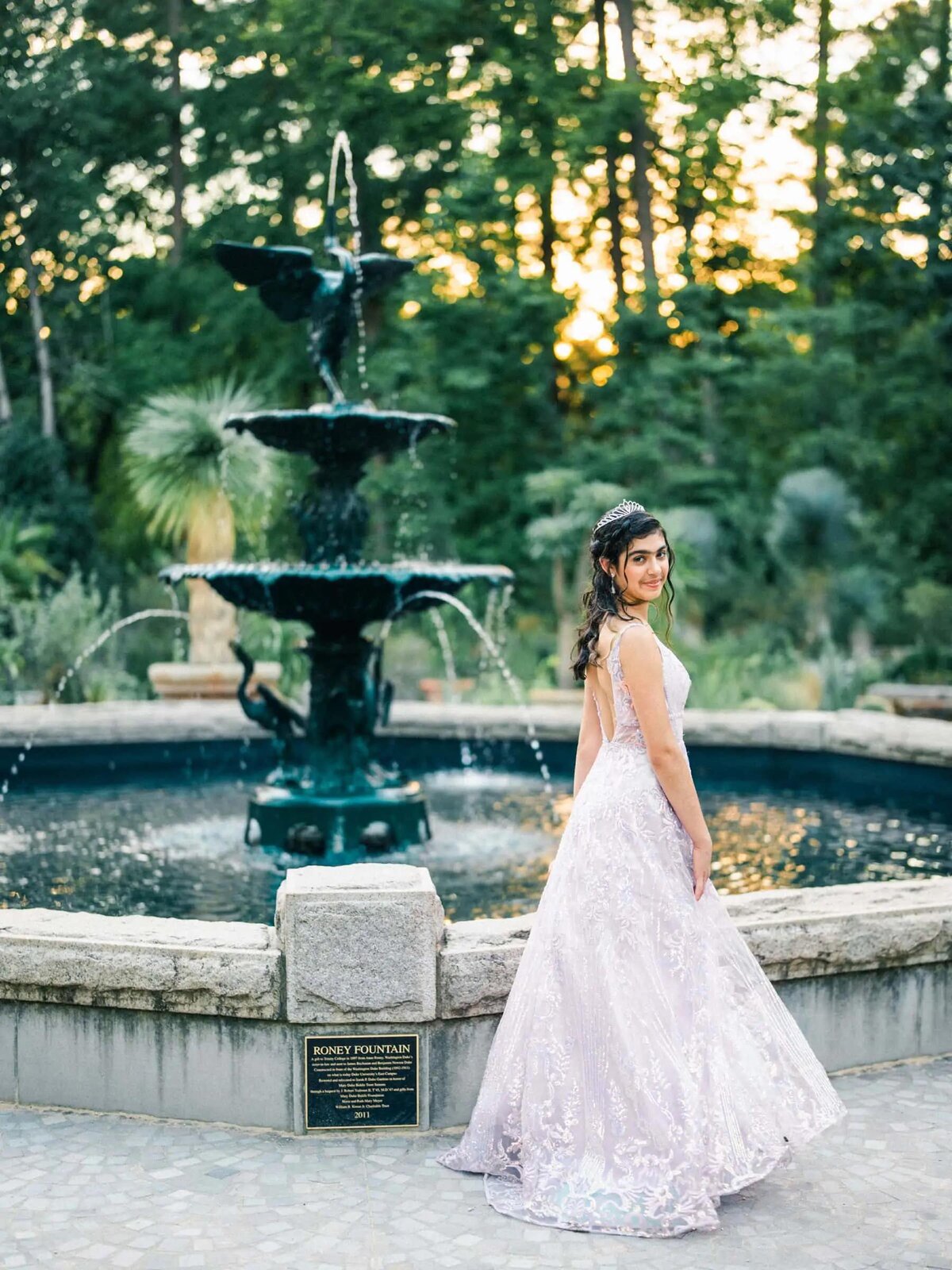 A girl in a gown standing in front of a fountain outside.