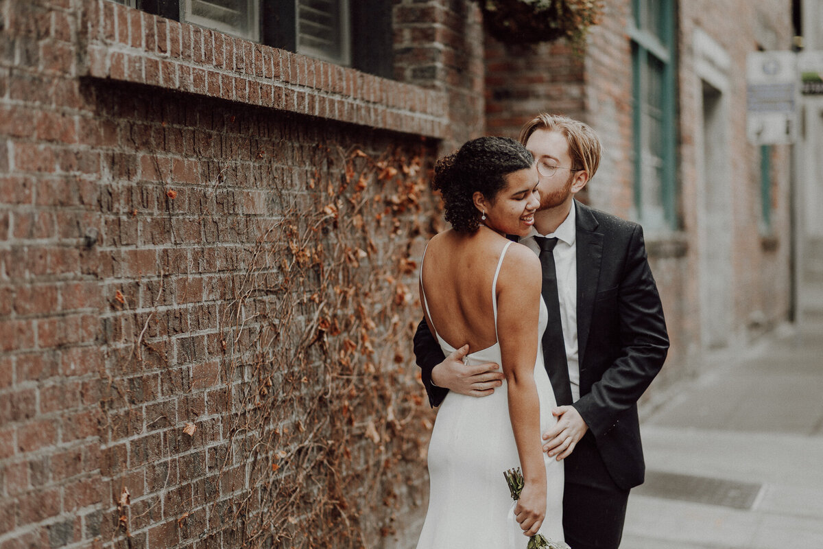 A groom kisses the cheek of his bride while they stand in an alley