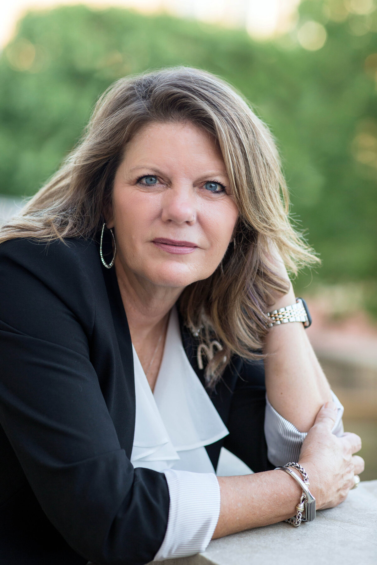 Woman is looking at the camera during an outdoor headshot photo session and is wearing a black suit jacket with white ruffled blouse and sliver hoop earrings