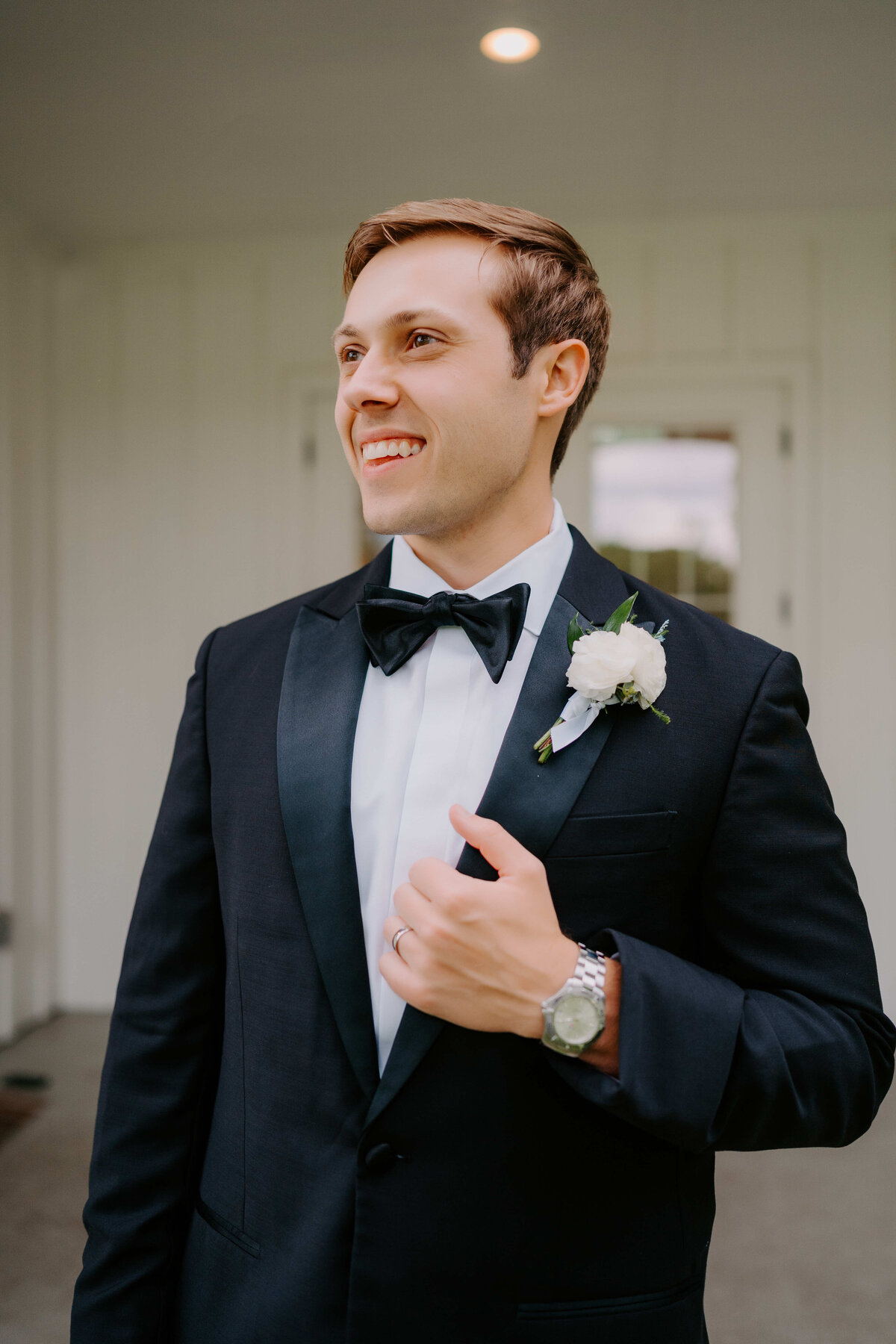 A smiling man in a tuxedo with a white boutonniere stands in front of a light-colored wall. He exudes happiness and confidence.