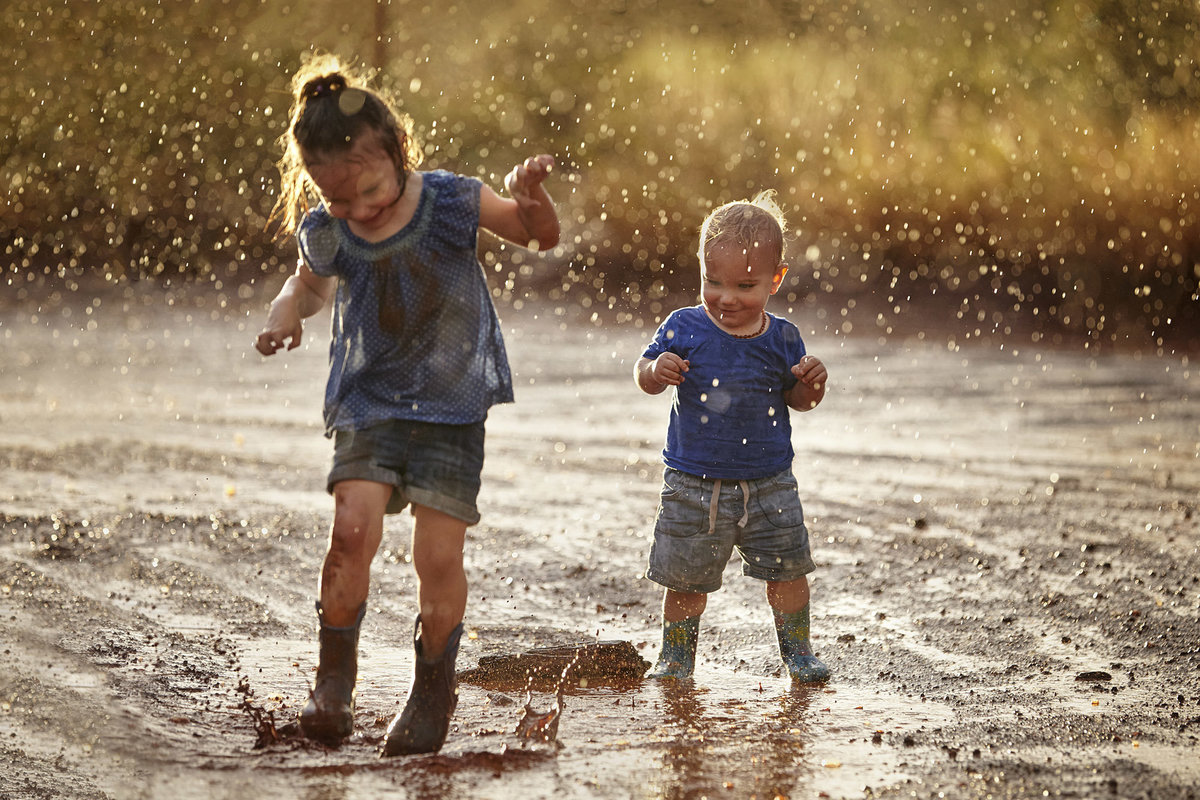 Young girl and boy in wellington boots splashing in puddles with rain falling down on them