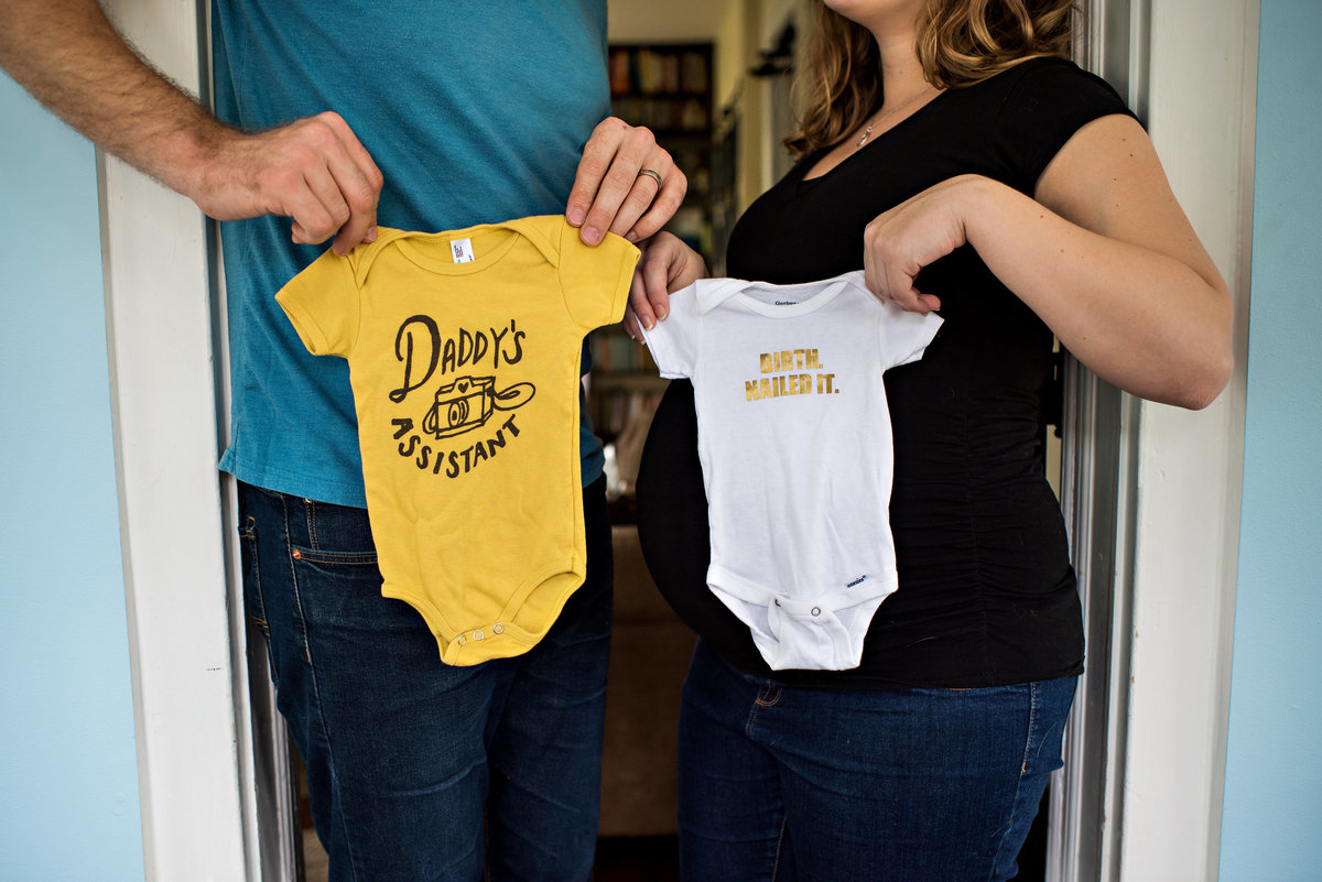 New parents hold up onesies for the new baby over the pregnant belly of the mother to be.