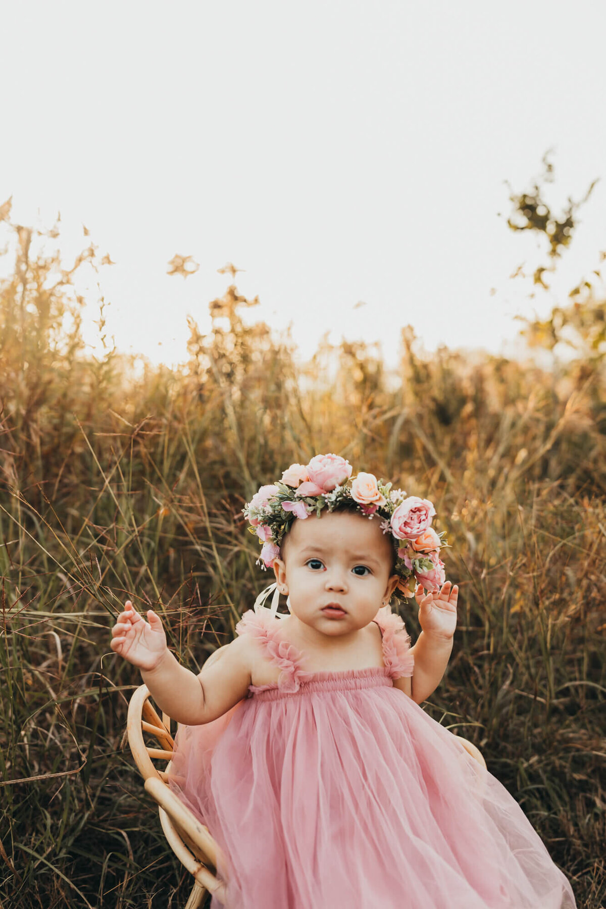 baby girl sits in wicker chair, with flower crown on her head and pink dress, in a field.