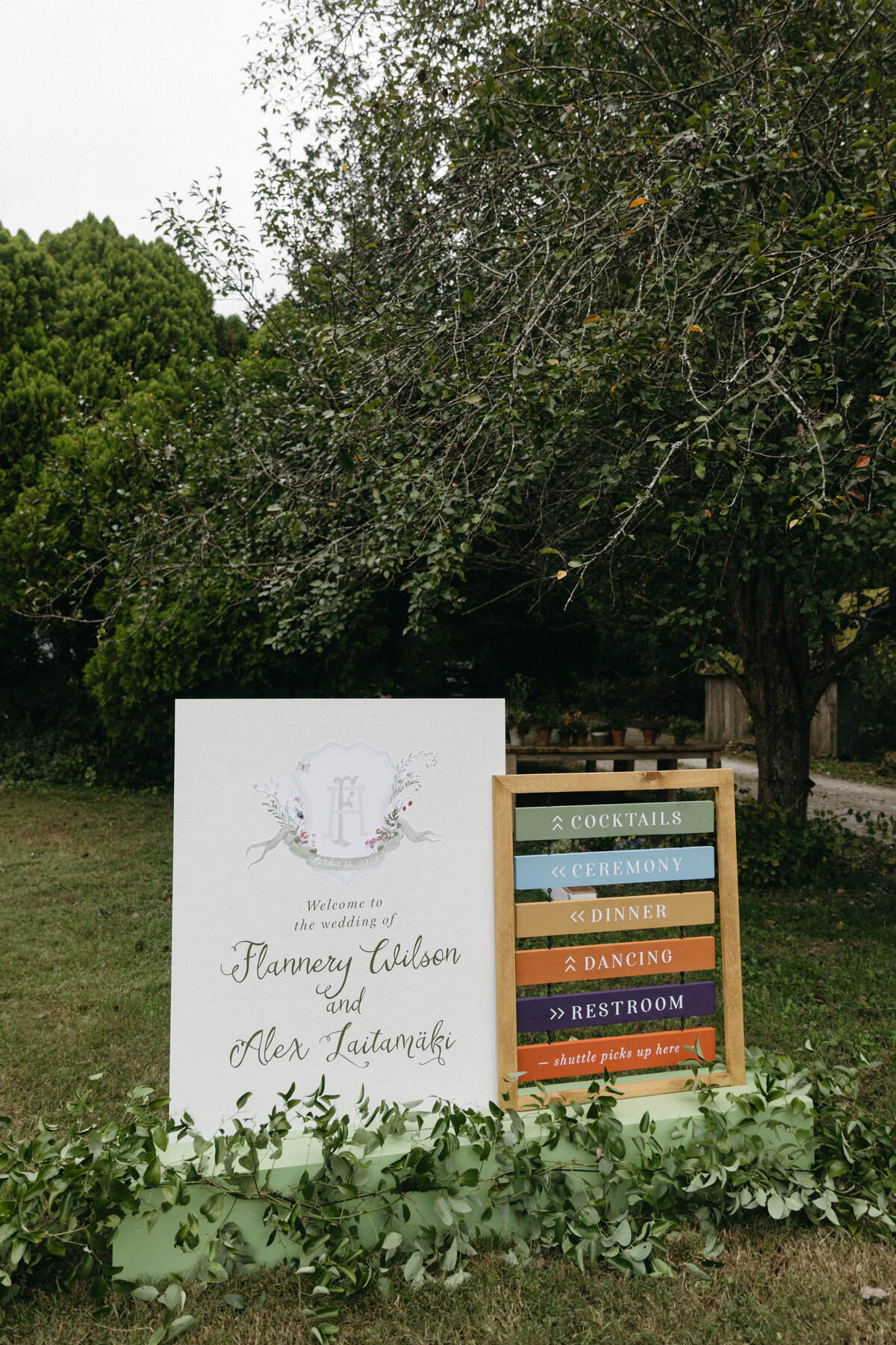 Outdoor wedding welcome display featuring a custom illustrated welcome sign and colorful wooden directional signage surrounded by fresh greenery—set at a garden-style Arkansas wedding designed with whimsical floral accents.