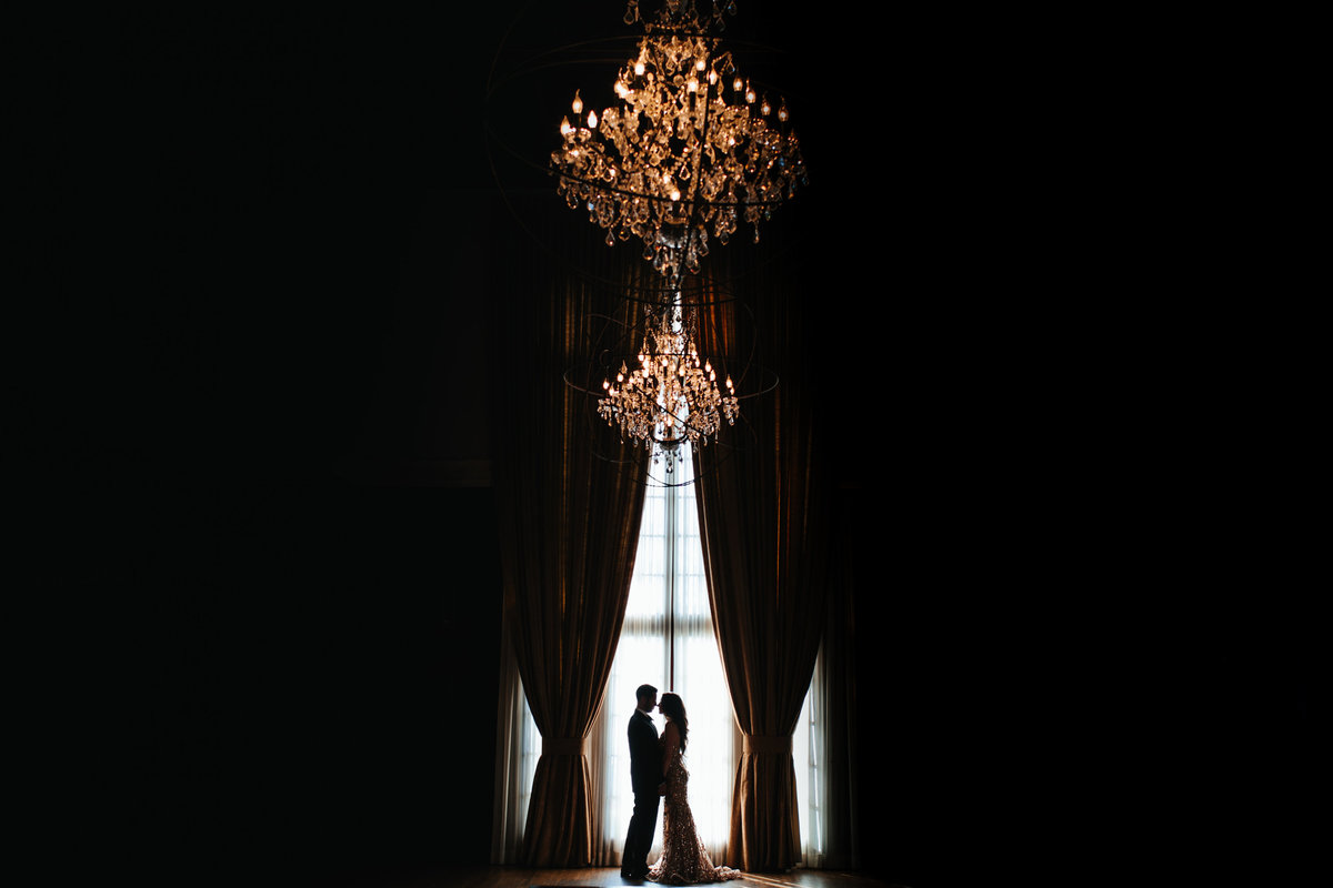 A dramatic photo of a couple against a giant window with large chandeliers over them