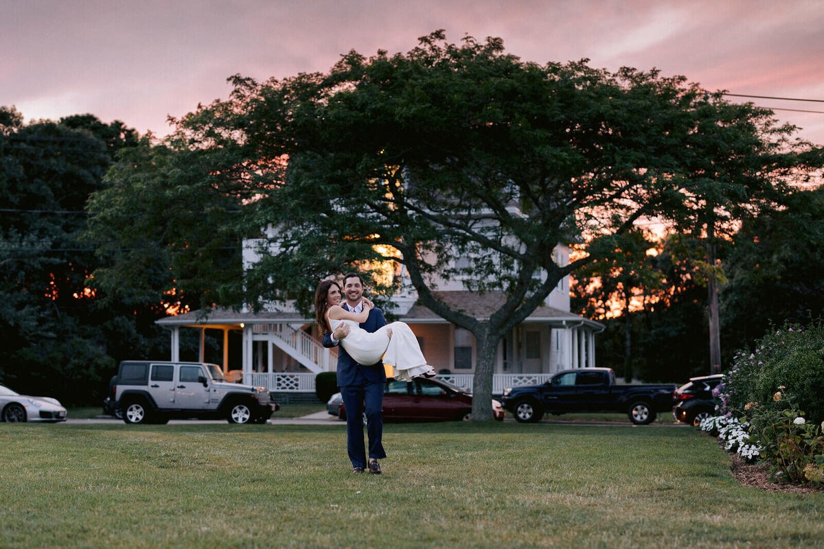 The groom is carrying the bride while standing outside a house at Cod Summer Tent, MA, with large trees in the background.