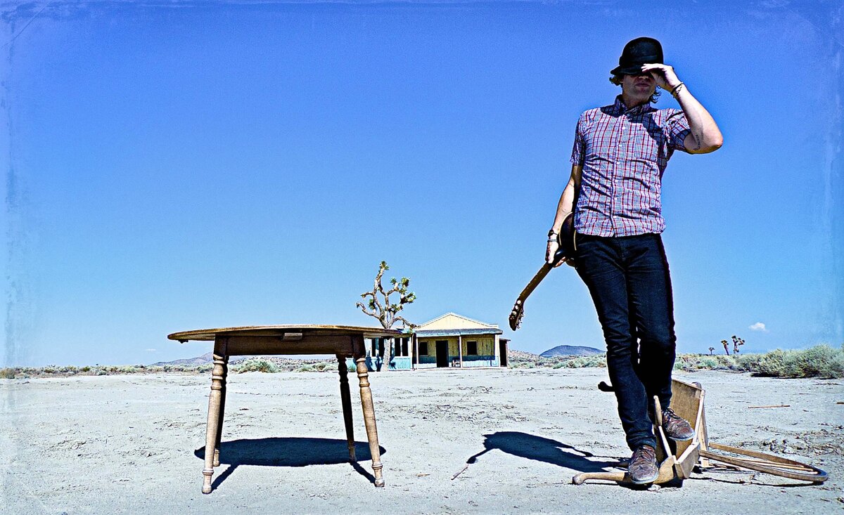 Male musician portrait Nico Stai wearing plaid shirt holding black fedora with guitar behind while standing upon broken wooden chair desert background