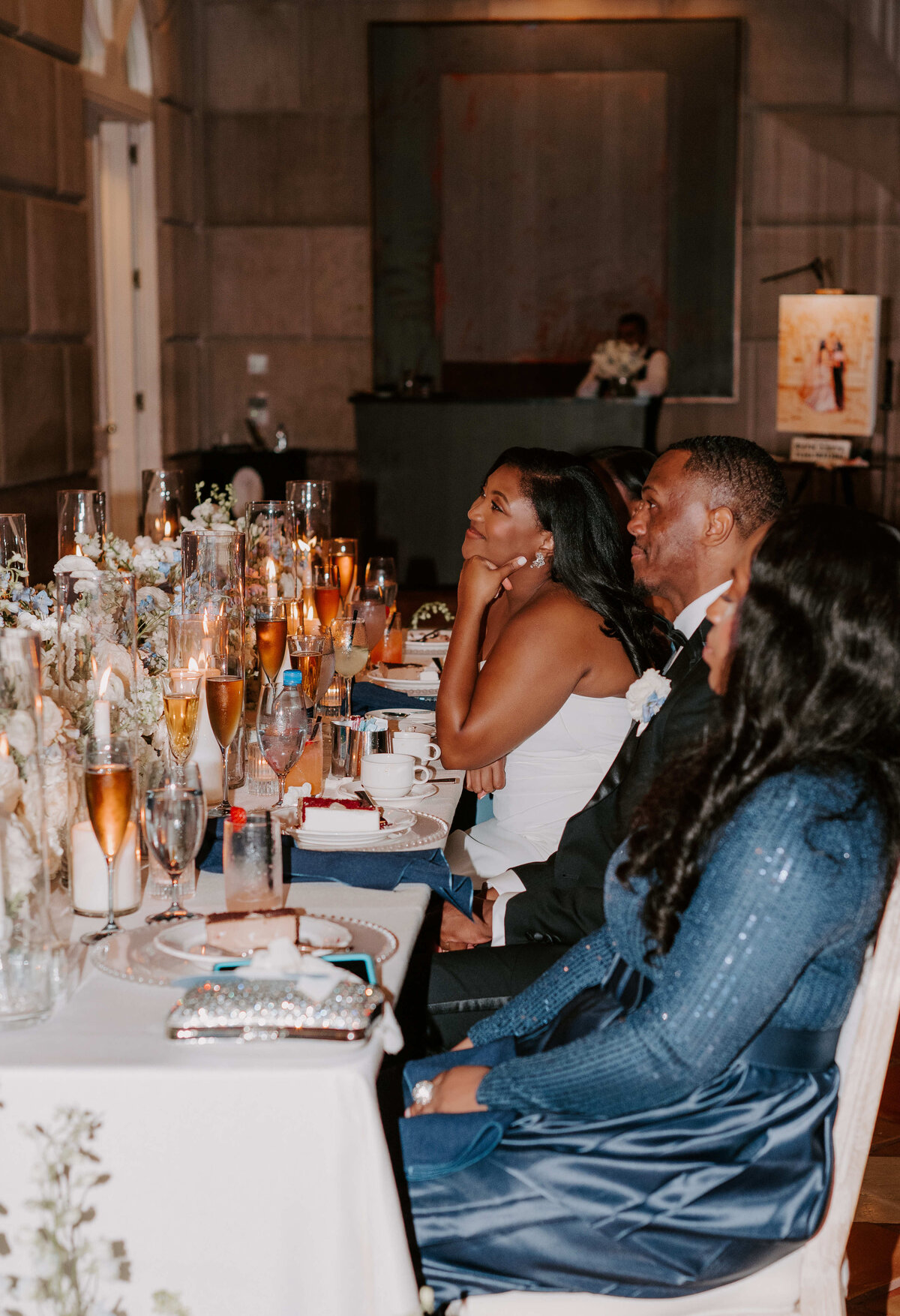 A couple sits at an elegantly decorated table with gold-rimmed glasses and flower arrangements, smiling and engaged, creating a warm, festive atmosphere.