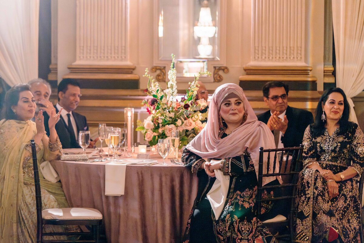 A group of people sit elegantly around a table decorated with flowers and candles in a formal setting. The attendees are dressed in formal attire and seem engaged, with some clapping and others smiling. The room is ornately decorated.