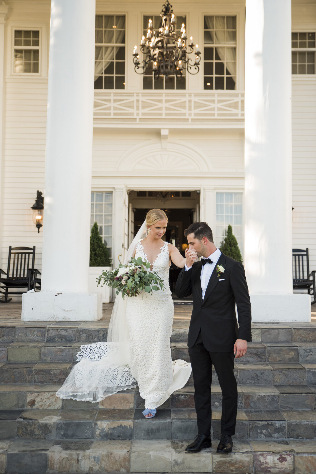 A groom leads his bride down the front steps of The Manor House and kisses her hand.
