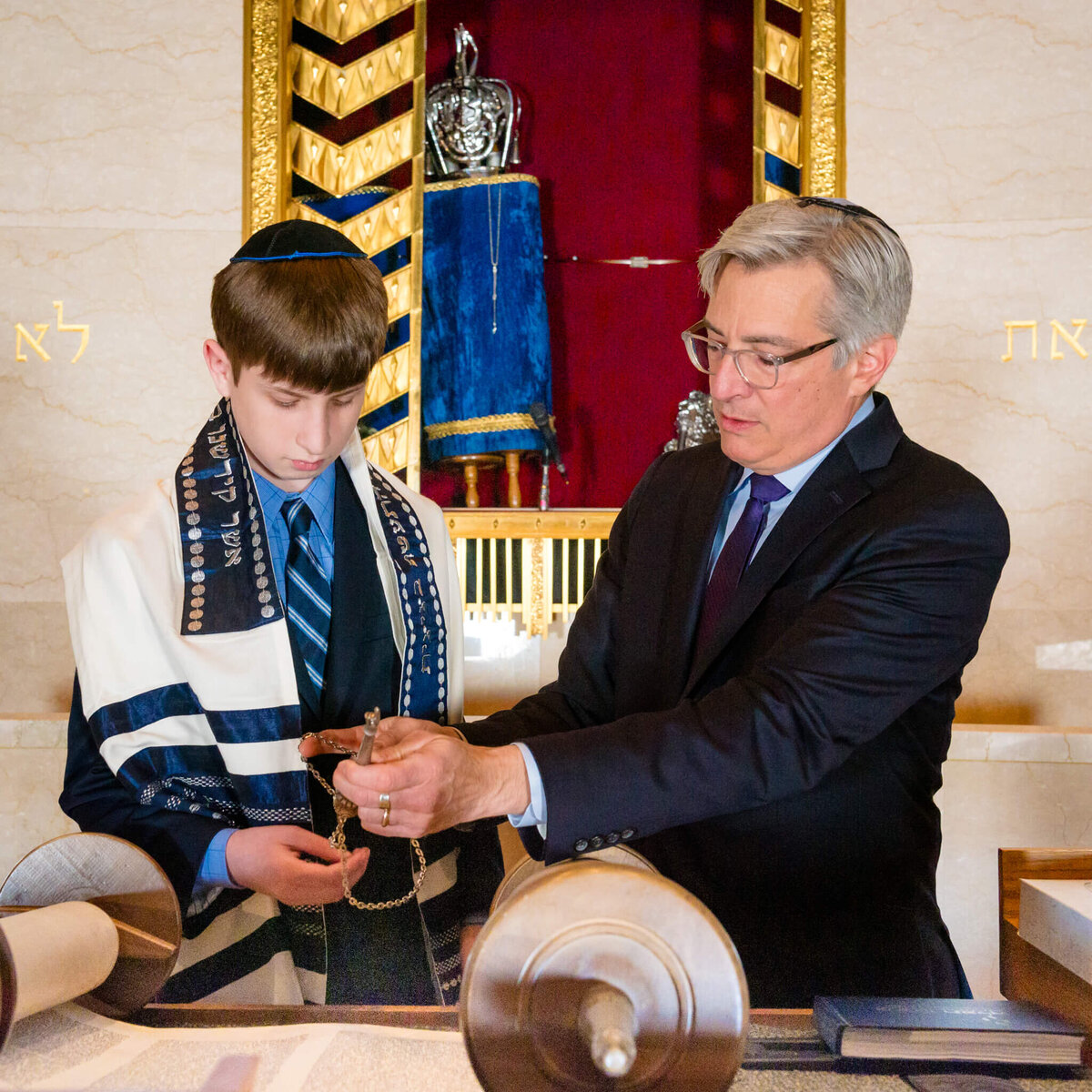 A rabbi helps a boy in a blue suit and tallit at the bimah