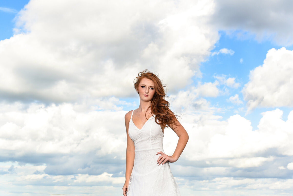 Richmond high school senior girl wearing white dress poses in the clouds at Yorktown Beach.