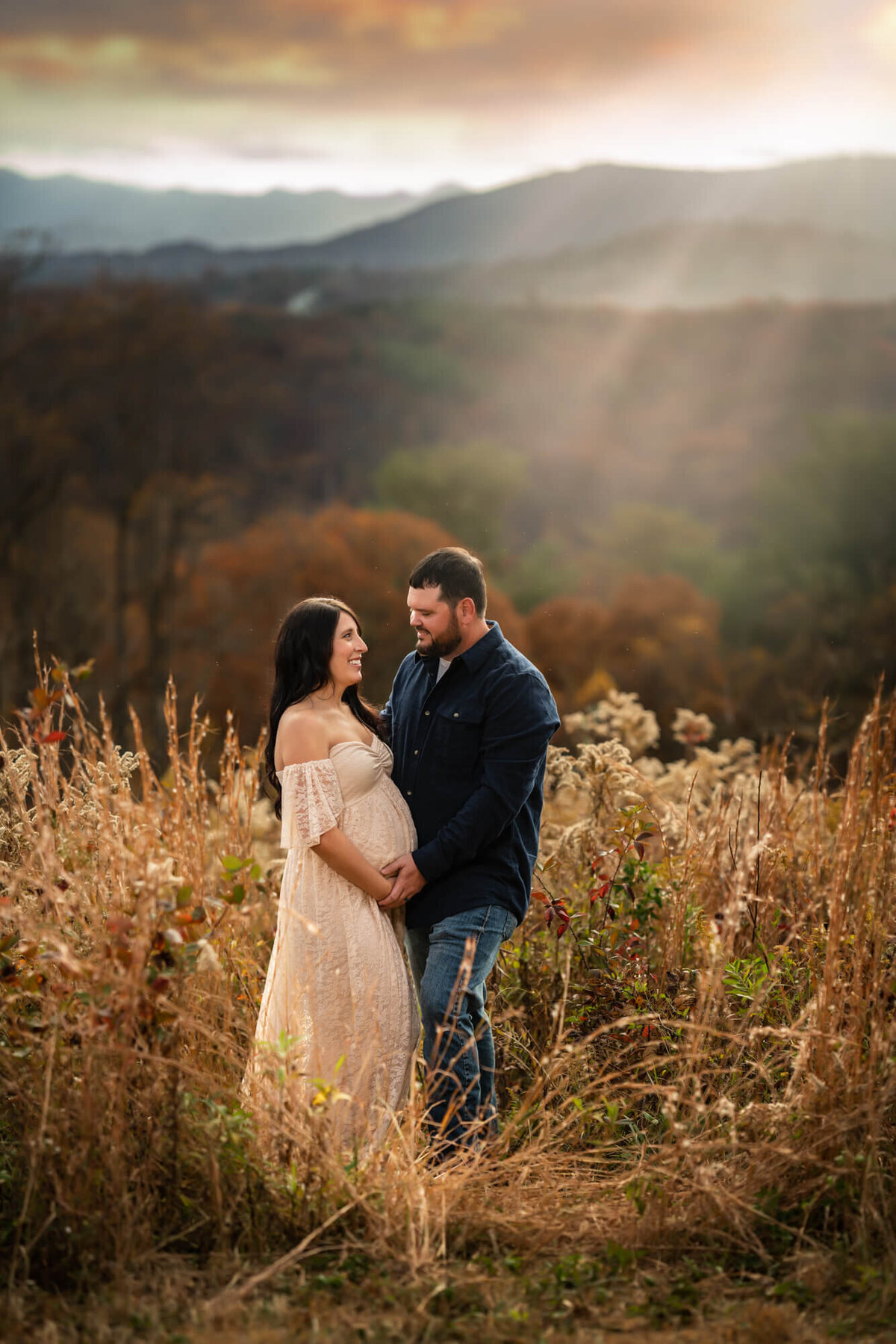 Sun rays pour over an expecting couple as they stand in tall grass
