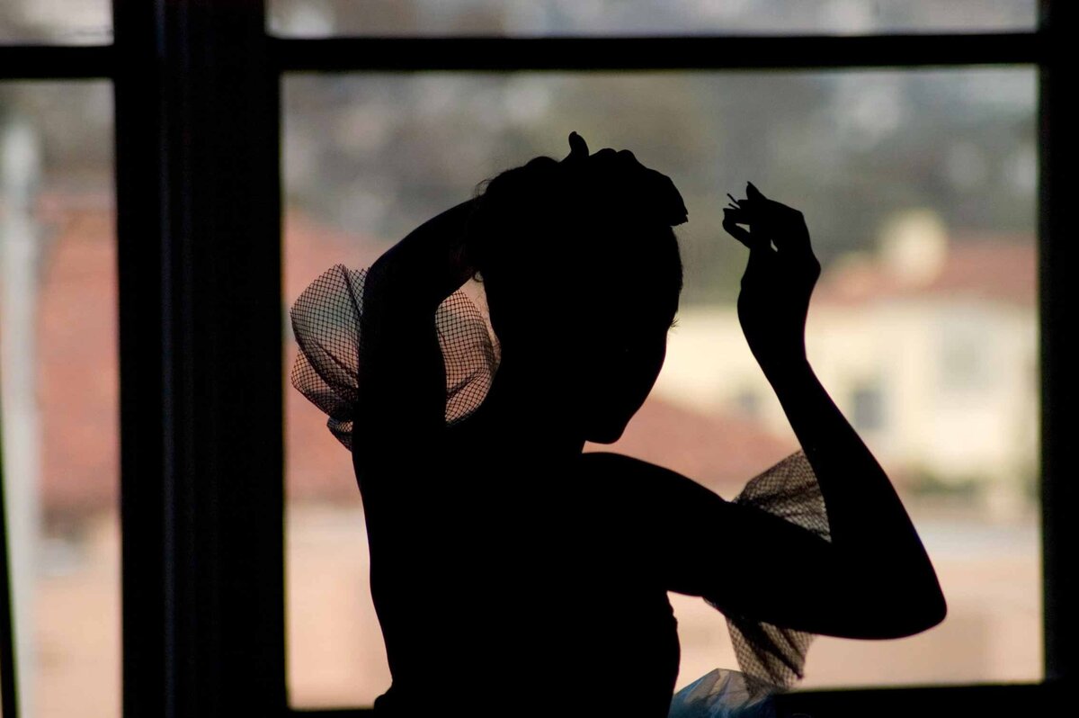 A ballet dancer put pin in her hair before performance. She is silhouetted against a window at liberty station in San Diego