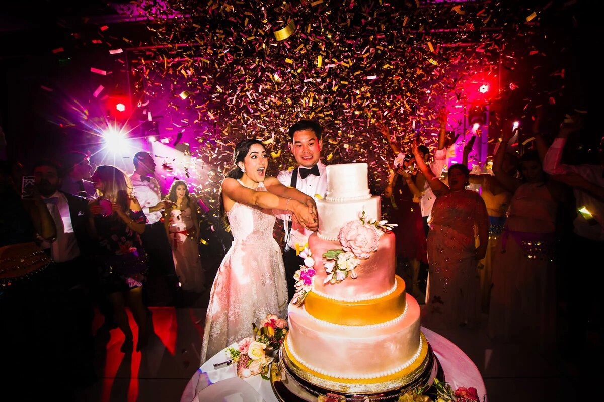 A bride and groom joyfully cut their wedding cake surrounded by vibrant lights and a shower of confetti